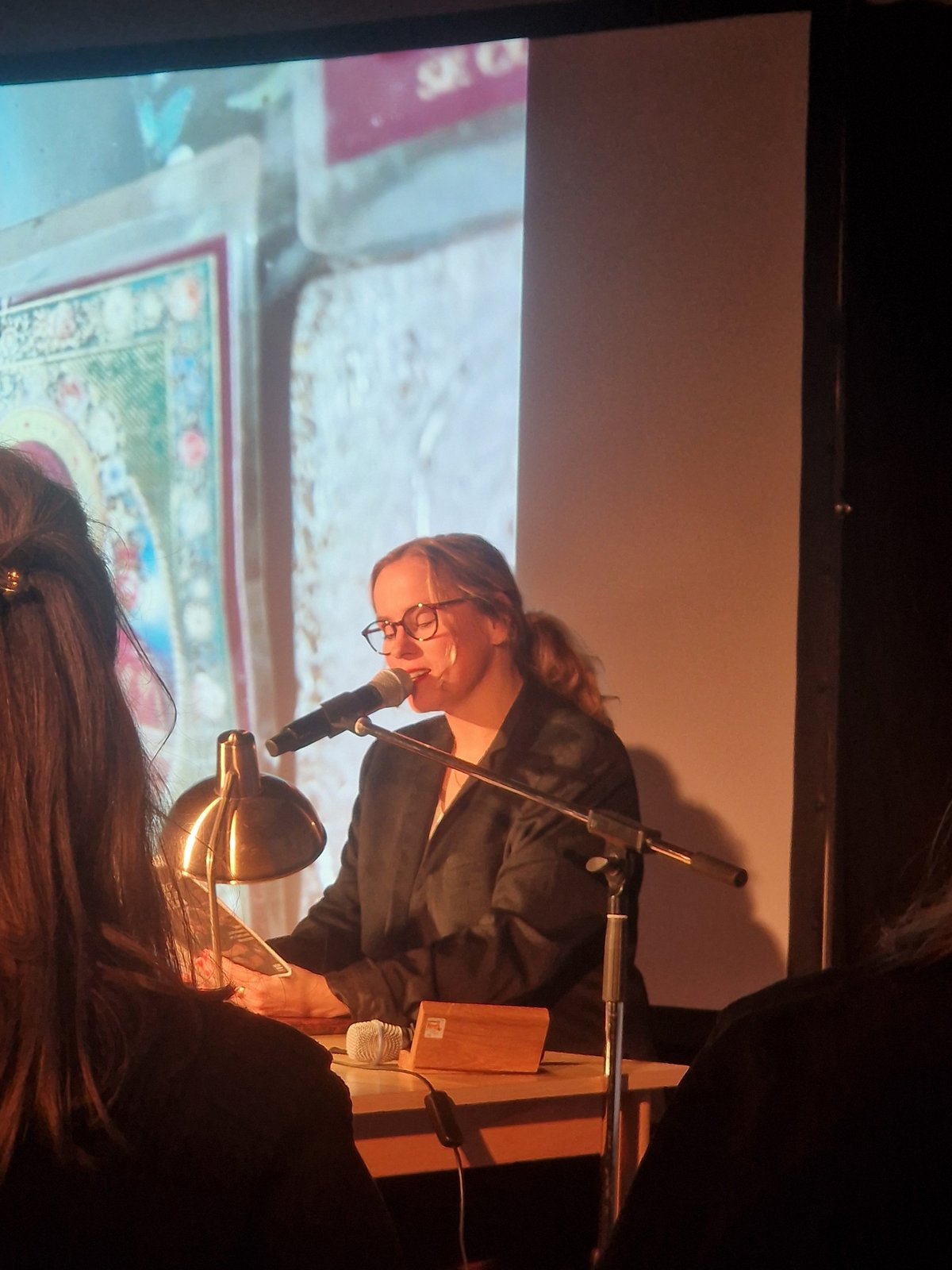 Sarah Brendel trägt ihren Text am Tisch vor eine Leinwand vor, auf der Bilder projeziert werden, passend zu ihren Texten.