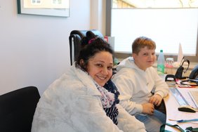 Ein Schüler sitzt mit einer Mitarbeiterin an ihrem Arbeitsplatz. Beide schauen lächelnd in die Kamera. 