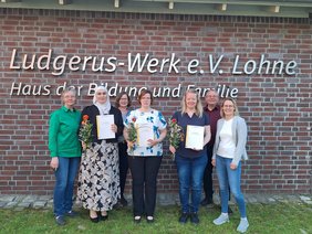 Die Gruppe der Teilnehmerinnen stehen mit den zuständigen Begleitern vor dem Logo an der Außenwand des Ludgerus-Werk. Die Teilnehmerinnen halten ihr Zertifikat und eine rot-orange Rose in den Händen. 