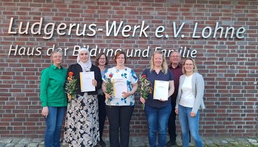 Die Gruppe der Teilnehmerinnen stehen mit den zuständigen Begleitern vor dem Logo an der Außenwand des Ludgerus-Werk. Die Teilnehmerinnen halten ihr Zertifikat und eine rot-orange Rose in den Händen. 