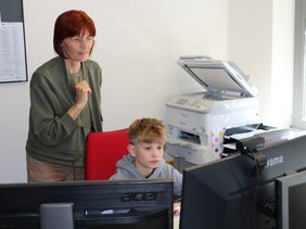 Ein Schüler sitzt am Arbeits-Computer im Büro. Eine Mitarbeiterin steht hinter ihm und zeigt ihm die Funktionsweise der Programme. 