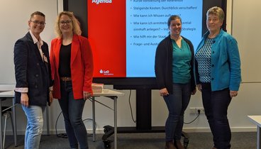 Vier Frauen stehen in jeweils in zwei Paaren neben einem Whiteboard. Sie schauen lächelnd in die Kamera. Auf dem Whiteboard ist die Agenda des Vortrags abgebildet.