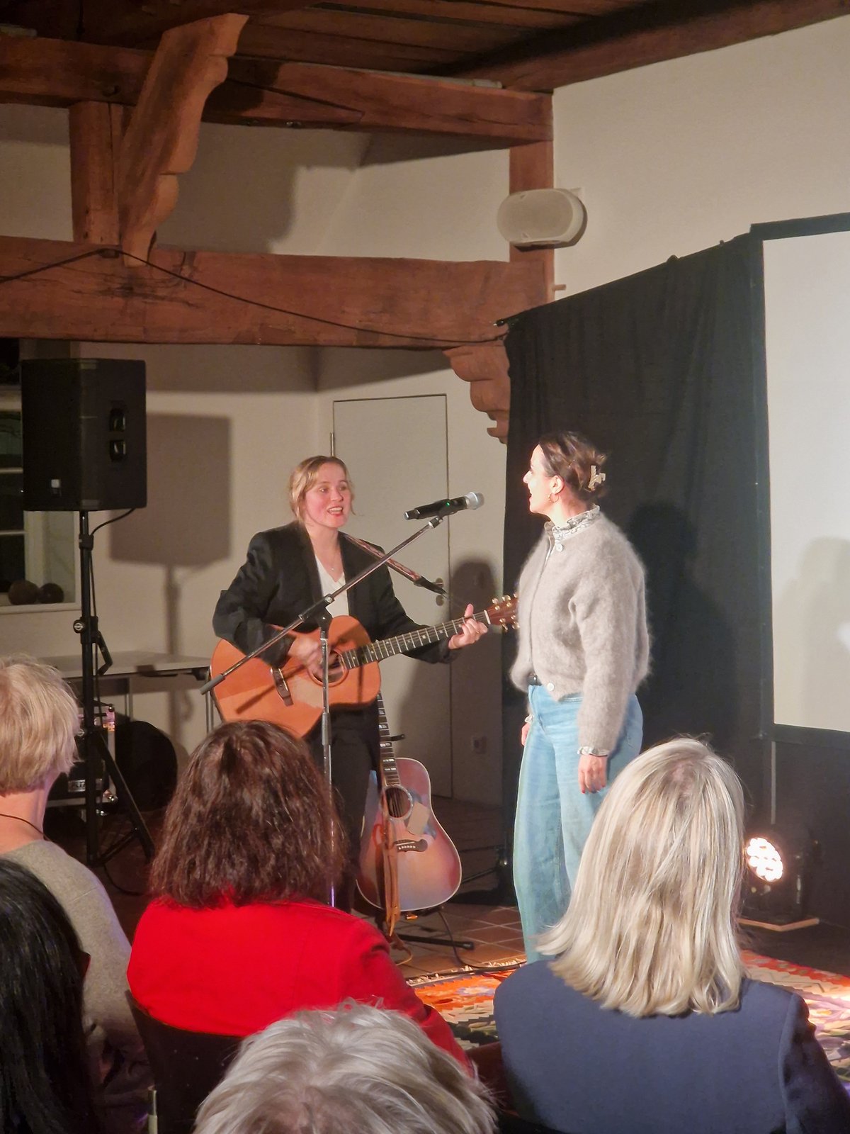 Sarah Brendel steht mit einer Freundin am Mikrofon und singt zusammen. Das Publikum ist von hinten zu sehen. 