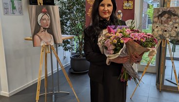 Künstlerin Veronika Fraile steht zwischen ihren Kunstwerken im Foyer des Ludgerus-Werks. Sie schaut lächeln in die Kamera und hält einen Blumenstrauß mit rosa Blumen in den Händen.
