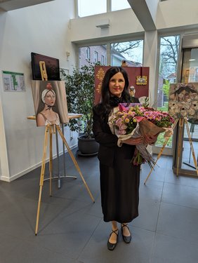 Künstlerin Veronika Fraile steht zwischen ihren Kunstwerken im Foyer des Ludgerus-Werks. Sie schaut lächeln in die Kamera und hält einen Blumenstrauß mit rosa Blumen in den Händen.