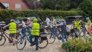 Eine Radfahrgruppe steht auf dem Parkplatz beim Ludgerus-Werk. Die Radfahrer tragen Helme und Warnwesten. 