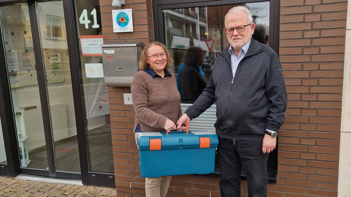 Zwei Mitarbeiter des Repair-Cafés stehen vor dem Eingang des Seniorentreffpunkts. Sie halten gemeinsam einen Werkzeugkoffer. 