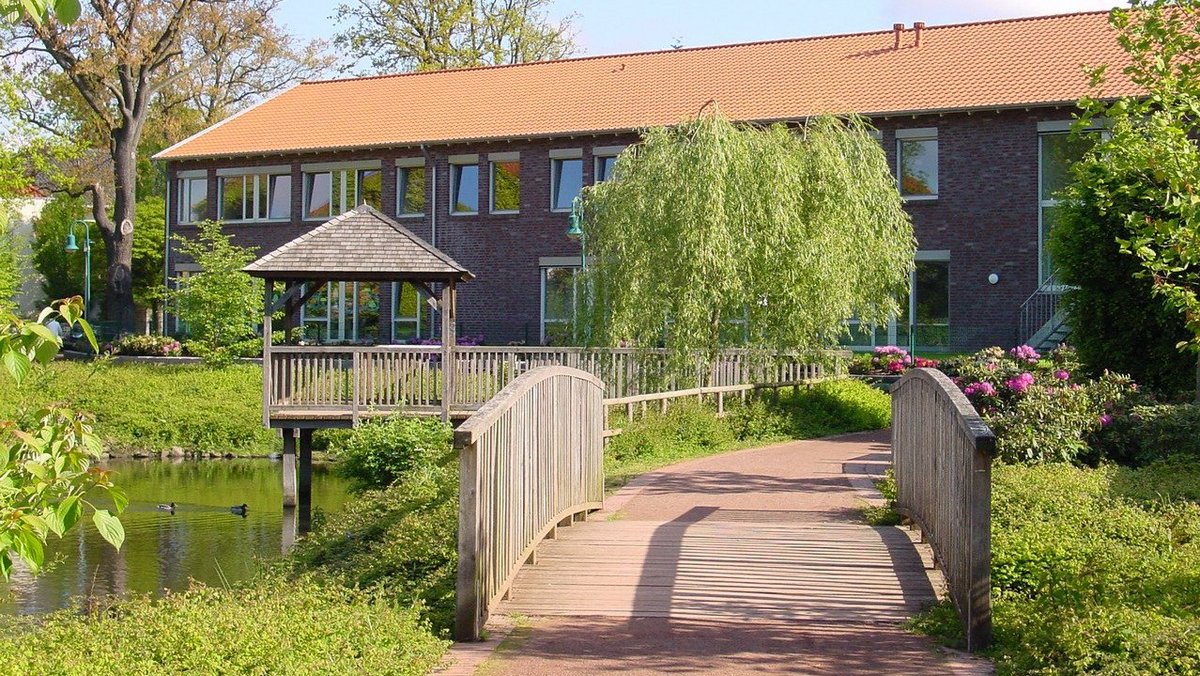 Man schaut auf den hinteren Teil des Ludgerus-Werk-Gebäudes. Im Vordergrund ist eine kleine Brücke. Links sieht man den Teich und einen Pavillon, der auf das Wasser reicht. Es ist ein sonniger Tag.