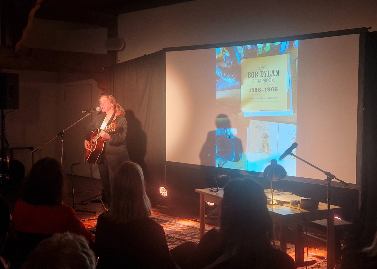 Die Künstlerin steht mit ihrer Gitarre am Mikrofon und singt. Im Hintergrund wird an die Leinwand eine Collage zum Musiker Bob Dylan projeziert. 