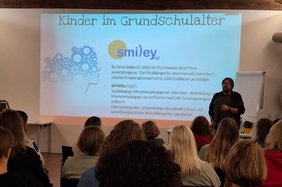 An eine weiße Wand ist eine PowerPointPräsentation projeziert. Daneben steht der Dozent Ralf Wilius. Im Vordergrund sin die Hinterköpfe der Zuhörenden zu sehen. 