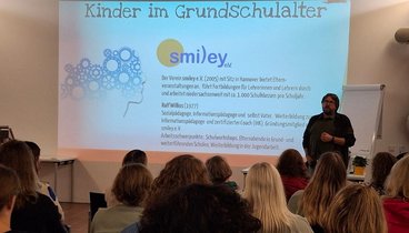 An eine weiße Wand ist eine PowerPointPräsentation projeziert. Daneben steht der Dozent Ralf Wilius. Im Vordergrund sin die Hinterköpfe der Zuhörenden zu sehen. 