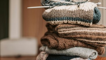 Ein Stapel gestrickter Objekte die zusammengelegt auf einem Tisch liegen. Obenauf liegt ein Knäuel Wolle mit Stricknadeln.