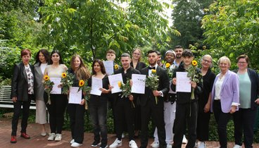 Die Gruppe von Teilnehmenden stehen zusammen mit den Lehrerinnen vor einer Reihe Büsche. Sie halten ihre Abschlusszeugnisse und eine Sonnenblume in den Händen