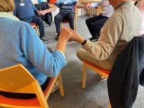 Die Teilnehmenden sitzen in einem Stuhlkreis und halten sich an den Händen. 