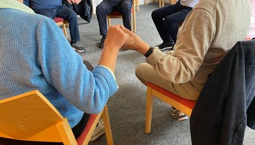 Die Teilnehmenden sitzen in einem Stuhlkreis und halten sich an den Händen. 