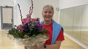 Bild von Doris Trenkamp in der Sporthalle im Ludgerus-Werk. Sie trägt ein rotes Shirt mit einem blauen Pullover über den Schultern. In den Händen hält sie einen großen Blumenstrauß. Die Blumen sind violett-pink und in Klarsichtfolie gepackt. 