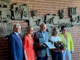 Eine Gruppe Leute stehen um den geehrten Werner Becker vor einer Ziegelwand mit Bildern daran. Werner Becker steht in der Mitte und hält eine Urkunde und eine Medaille in einem Kästchen in der Hand.