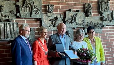 Eine Gruppe Leute stehen um den geehrten Werner Becker vor einer Ziegelwand mit Bildern daran. Werner Becker steht in der Mitte und hält eine Urkunde und eine Medaille in einem Kästchen in der Hand.