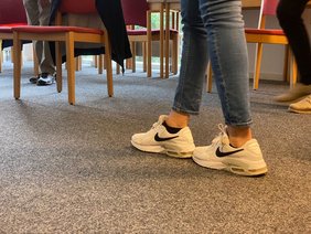 Die Kursleitung macht eine Übung vor. Füße werden direkt voreinander für eine Balanceübung gesetzt. 
