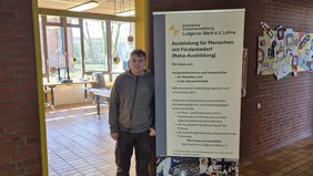 Es ist der Flur einer Schule zu sehen. Auf der linken Seite ist ein Türdurchgang. Man sieht einen Werkraum. Vor der Tür steht ein junger Mann neben einem Roll-Up. Auf dem Roll-Up stehen Informationen zur Berufsorientierung.