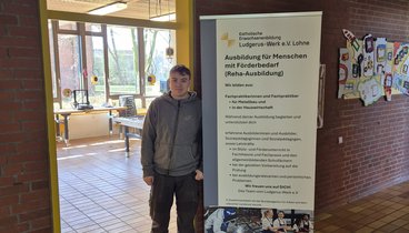 Es ist der Flur einer Schule zu sehen. Auf der linken Seite ist ein Türdurchgang. Man sieht einen Werkraum. Vor der Tür steht ein junger Mann neben einem Roll-Up. Auf dem Roll-Up stehen Informationen zur Berufsorientierung.