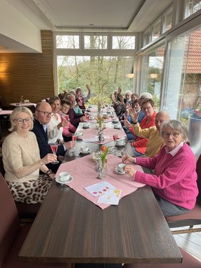 Eine Gruppe Menschen sitzt an einem langen Tisch. Sie schauen in die Kamera. EInige haben ein Sektglas in der Hand.