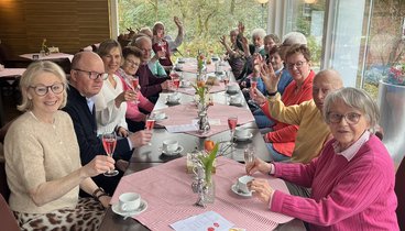Eine Gruppe Menschen sitzt an einem langen Tisch. Sie schauen in die Kamera. EInige haben ein Sektglas in der Hand.