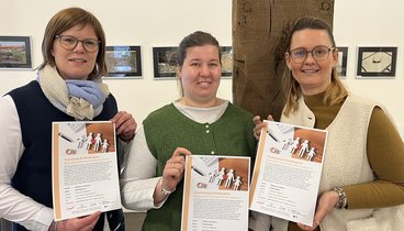 Drei Frauen halten das Plakat zum Vortrag in der Hand. Sie schauen in die Kamera. 