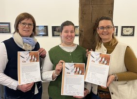 Drei Frauen halten das Plakat zum Vortrag in der Hand. Sie schauen in die Kamera. 