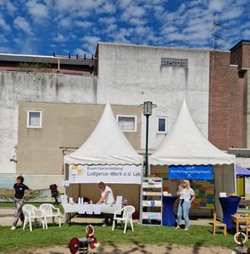 Es sind zwei Pavillons zu sehen. Einer ist der Ausstellungspavillon des Ludgerus-Werks, der andere der des Kindertagespflegebüros. Vor den Pavillons stehen Tische und Stühle. Drei Mitarbeiter sind an den Ständen beschäftigt.. Es ist gutes Wetter und blauer Himmel.  