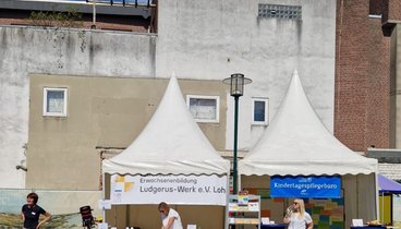 Es sind zwei Pavillons zu sehen. Einer ist der Ausstellungspavillon des Ludgerus-Werks, der andere der des Kindertagespflegebüros. Vor den Pavillons stehen Tische und Stühle. Drei Mitarbeiter sind an den Ständen beschäftigt.. Es ist gutes Wetter und blauer Himmel.  