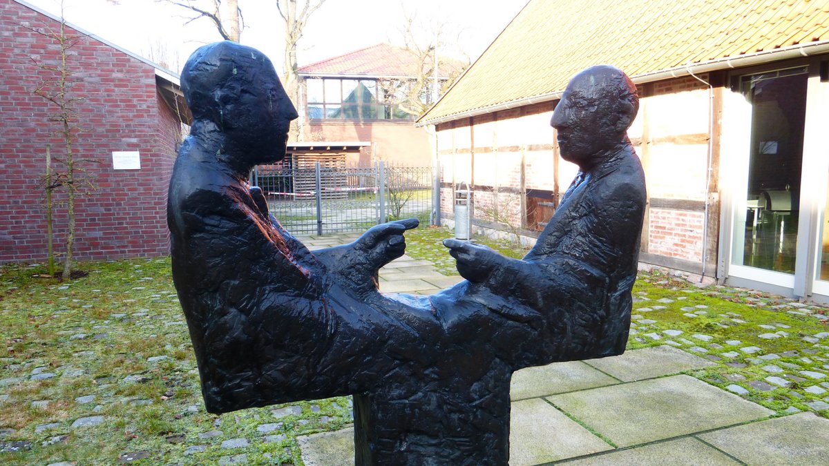 Eine eiserne Skulptur von zwei stilisierten Männern. Sie sehen sich an. Die Männer haben jeweils eine Hand erhoben. Es sieht aus, als ob sie diskutieren. 