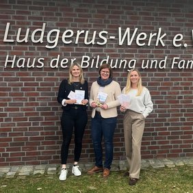 Drei Frauen stehen vor dem Logo an der Außenwand des Ludgerus-Werk e.V. Lohne. 