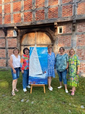 Fünf Künstlerinnen der Ausstellung Meer&Mehr stehen um ein Kunstwerk herum. Sie stehen draußen vor der Hofstelle des Ludgerus-Werks. Das Kunstwerk zeigt ein Segelboot auf dem Wasser. Das Segel des Bootes ist aus Stoff und ragt über den Rahmen hinaus. 
