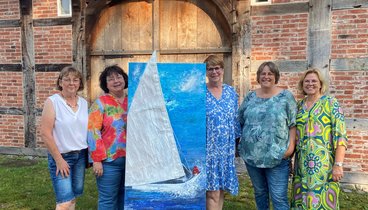 Fünf Künstlerinnen der Ausstellung Meer&Mehr stehen um ein Kunstwerk herum. Sie stehen draußen vor der Hofstelle des Ludgerus-Werks. Das Kunstwerk zeigt ein Segelboot auf dem Wasser. Das Segel des Bootes ist aus Stoff und ragt über den Rahmen hinaus. 