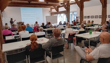 Die Hofstelle des Ludgerus-Werk. Die Teilnehmenden schauen nach vorne auf eine Leinwand, wo der Vortrag projiziert wird. Die Teilnehmenden sitzen an Tischen.  