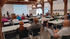 Die Hofstelle des Ludgerus-Werk. Die Teilnehmenden schauen nach vorne auf eine Leinwand, wo der Vortrag projiziert wird. Die Teilnehmenden sitzen an Tischen.  