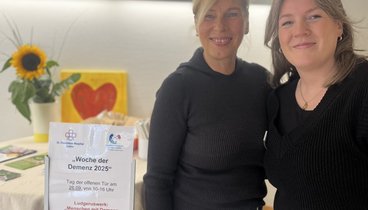Marika Görtz-Ochotzki und Sabrina Diekhaus stehen an einem Stehtisch mit freundlicher Blumendekoration und Informationsblättern zu Menschen mit Demenz bewegen.