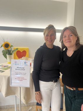 Marika Görtz-Ochotzki und Sabrina Diekhaus stehen an einem Stehtisch mit freundlicher Blumendekoration und Informationsblättern zu Menschen mit Demenz bewegen.