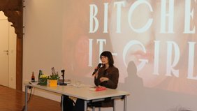 Autorin Rebekka Endler sitzt am Lesungstisch vor einer Landwand mit dem Titel ihres Buches projeziert. 