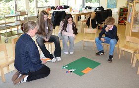 Betreuerin Sabrina Diekhaus vom Ludgerus-Werk sitzt mit den Teilnehmeden in einem Stuhlkreis um ein Plakat. Das grüne Plakat ist mit "Pilates" beschriftet. 