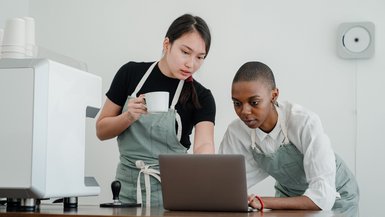 Zwei junge Frauen stehen vor einer Theke. Sie beugen sich über einen Laptop, der auf der Theke steht.