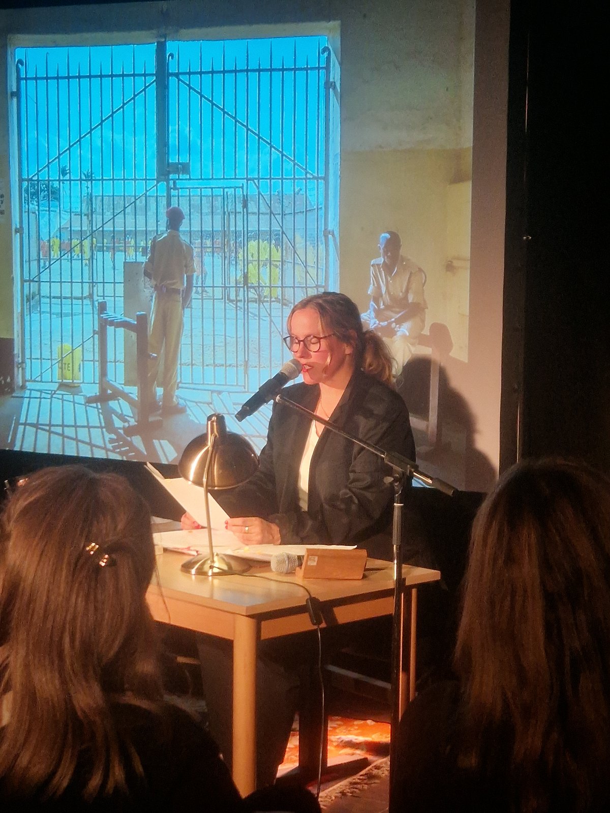 Sarah Brendel lesenderweise vor der Leinwand auf der ein Bild aus einem Gefängnis in Afrika projeziert wird. 