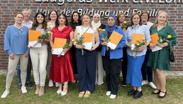 Eine Gruppe aus 15 Frauen stehen in zwei Reihen vor dem Logo an der Außenwand des Ludgerus-Werks. Die Absolventinnen halten eine Urkunde und eine weiße Rose in den Händen. Alle posieren lächelnd für die Kamera. 