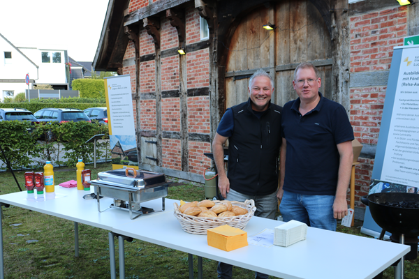Zwei Kollegen stehen an dem Tisch mit den Grills und der Bratwurststation.